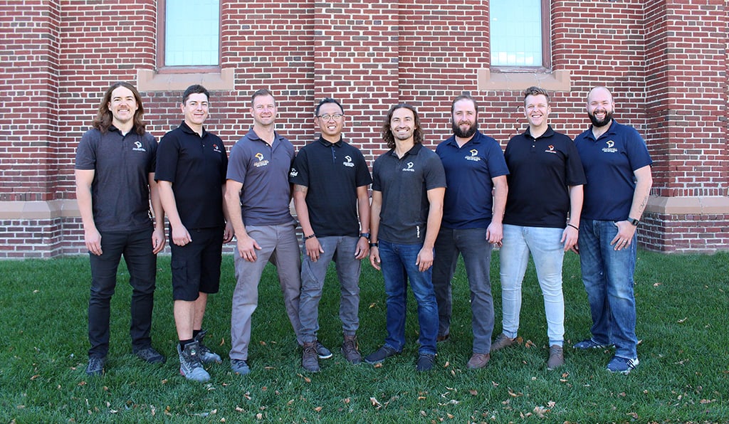 8 team members of collaborative construction standing in front of brick building on grass