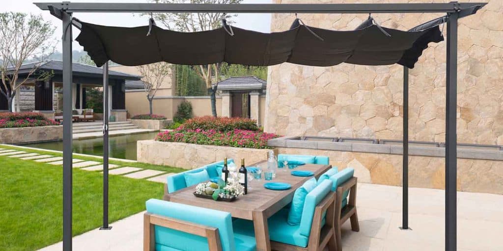 steel pergola in a yard on a rock patio with a set table and bright blue cushioned outdoor chairs