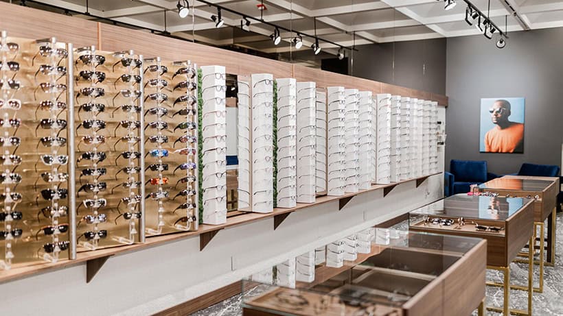 Modern optical retail store with wall-mounted white display towers showcasing various eyeglass frames, alongside wooden racks holding sunglasses. Sleek wooden display counters with glass tops line the space.